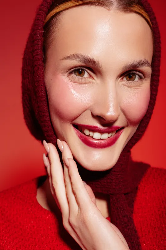 Smiling woman with clear skin wearing a red scarf and sweater poses against a red background. Close-up portrait with natural makeup and joyful expression. Concept of beauty and fashion.