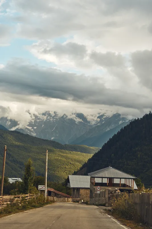 Mountain village road to rustic house and valley under dramatic clouds, green forest slopes and distant peaks, rural landscape with authenticity inclusivity and soft light