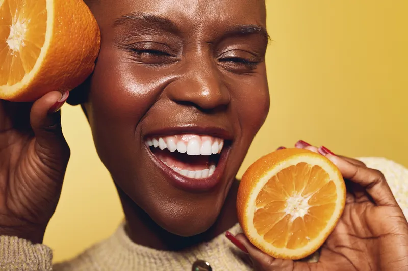 Close-up of a smiling dark skinned African woman holding two orange halves near her face on yellow background. Concept of healthy lifestyle, fresh fruit, happiness, and natural beauty.