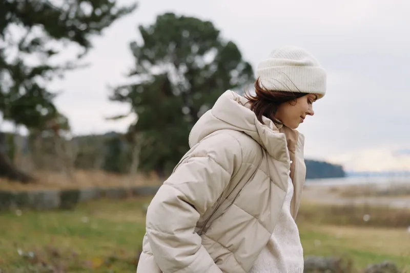 Woman in a beige puffer coat and knit beanie walking outdoors along a windy field with trees in the background, capturing a calm cold weather mood and simple natural light