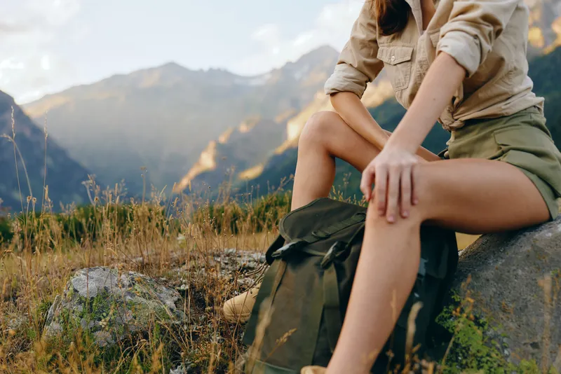 Cheerful woman outdoors, resting on a rock in a mountain landscape, wearing casual hiking clothes, sunlit moment, relaxed posture, natural tones, expansive scenery. Calm.