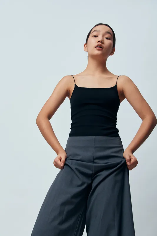 Young woman standing with hands in pockets wearing black tank top and wide gray pants against a plain light background. Fashion, casual style and confident pose in studio portrait.