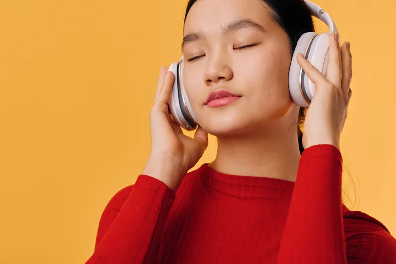 Woman with closed eyes listening to music on wireless headphones with relaxed expression in red sweater against orange background. Portrait of calm female enjoying sound.
