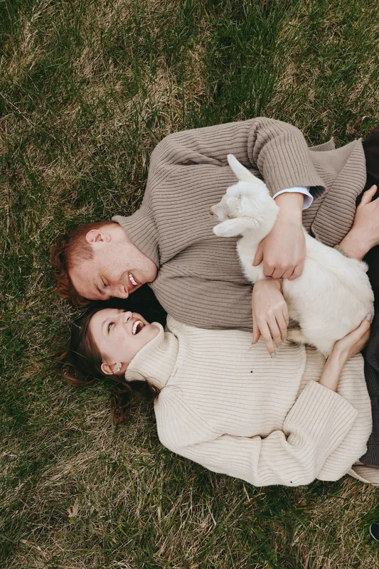 A couple lies on a grassy field with a white goat kid resting between them, capturing a warm, intimate lifestyle moment in a natural outdoor setting. Tender affection, relaxation, and rural
