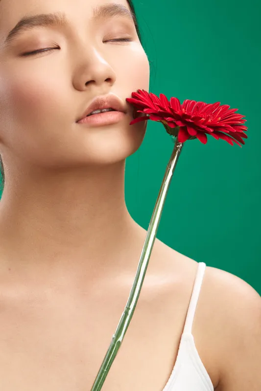 asian woman appears serene in a minimalist portrait, resting beside a red flower against a vivid green background, conveying calm, focus, and graceful contemplation.