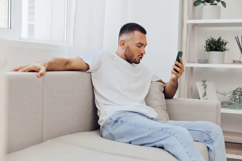 Focused man using smartphone while sitting on a stylish sofa, showcasing a modern living space with natural light and a calm atmosphere. Ideal for lifestyle themes.