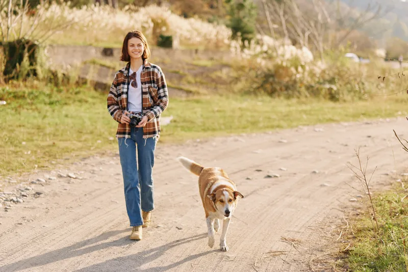 Woman walking with a dog on a sunny countryside path, wearing a plaid shirt, jeans and boots, enjoying nature, fresh air and peaceful scenery along an open field trail in daylight.