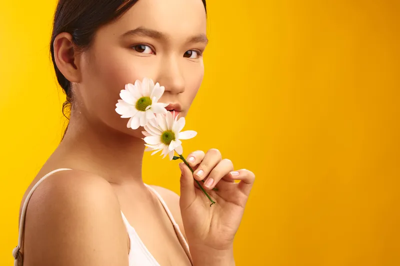 Asian woman holds a white flower near her lips against a bright yellow background, soft lighting enhances natural features, creating a fresh, confident portrait.