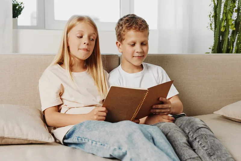 Two children sitting on a sofa reading a book together in a bright living room, sharing a calm and focused moment.