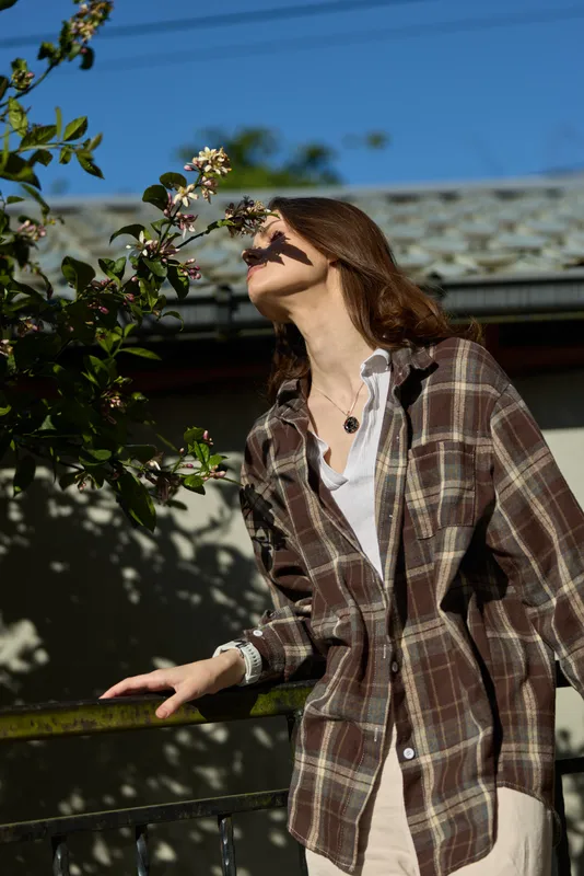 A woman in a plaid shirt stands on a sunny balcony, gazing upward beside blooming branches, creating a casual lifestyle moment that conveys relaxed outdoor leisure