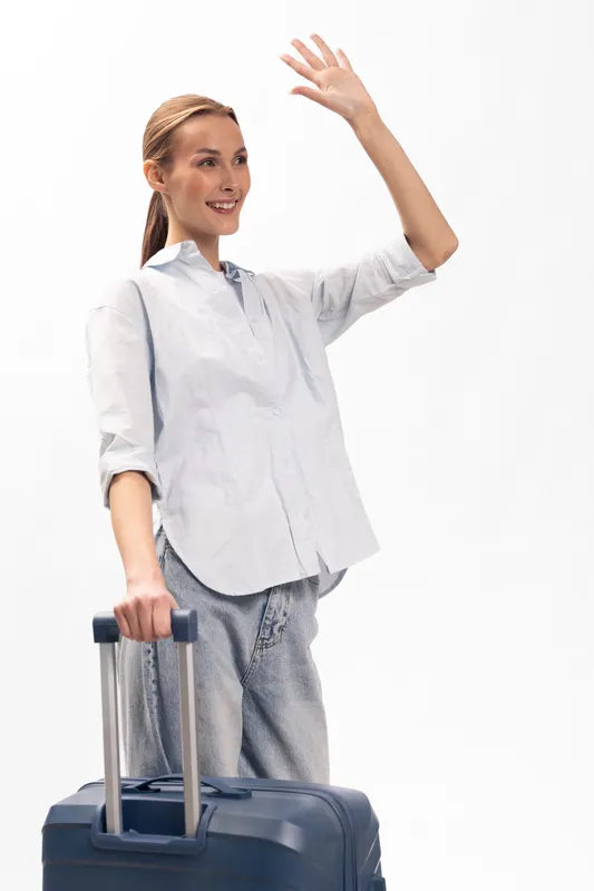 Smiling woman with ponytail holds suitcase handle and waves hand. She wears casual shirt and pants, isolated on white background. Concept of travel, journey, and friendly greeting.