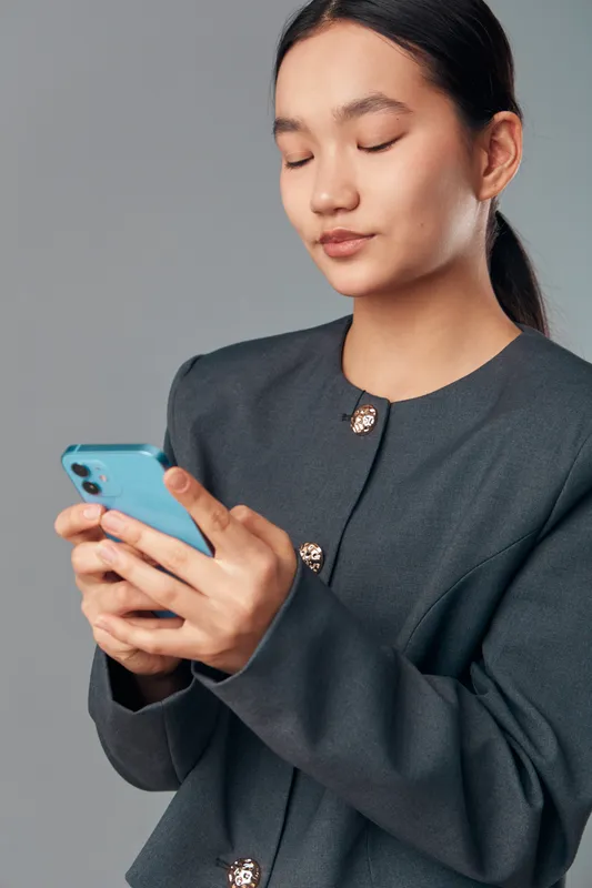 Young woman holding smartphone and texting with focused expression in studio, wearing dark blazer with buttons and standing against plain background with smartphone use concept.