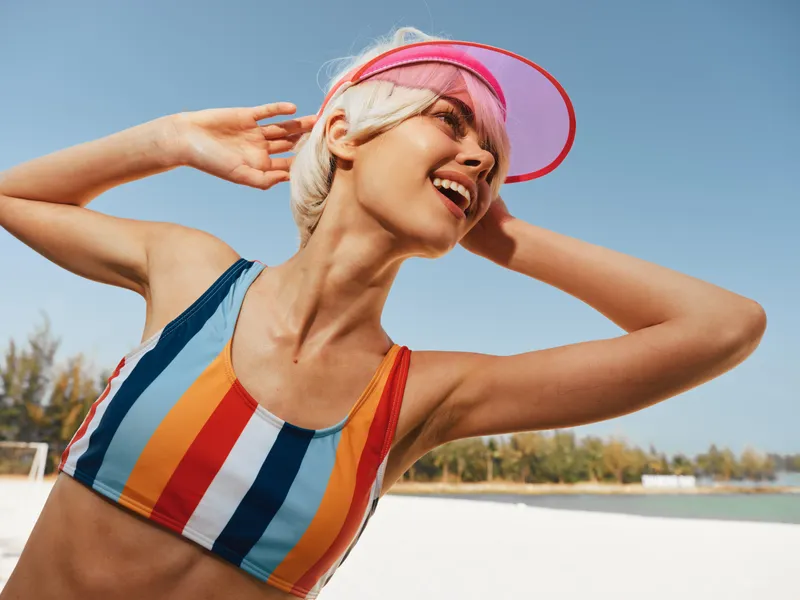Joyful young woman at the beach wearing a colorful striped swimsuit and pink visor, posing with arms raised, sunlit sky, carefree summer vibes.