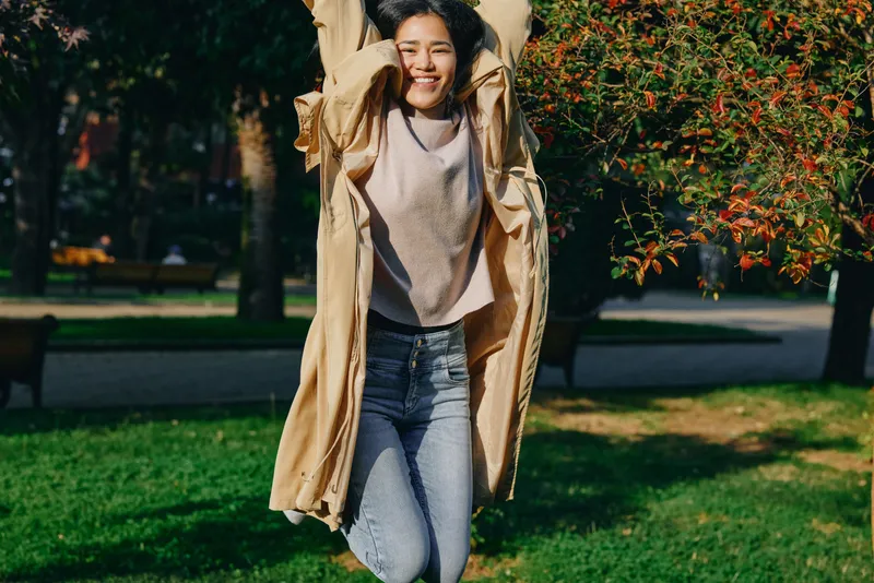 Woman jumping in a lightweight coat in an outdoor autumn park, showcasing motion, happiness, and cheerful style as leaves flutter and sunlight warms the candid scene