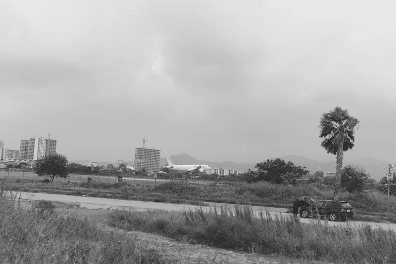 Monochrome landscape with distant cityscape on the horizon, a lone car on a dirt road, tall palm, open field, and moody sky; contrasts of urban proximity and rural calm, suitable for travel,