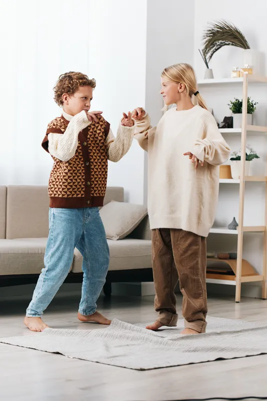 Two playful children dance indoors, wearing cozy sweaters and holding hands, as a warm livingroom scene glows with natural light and cheerful energy.