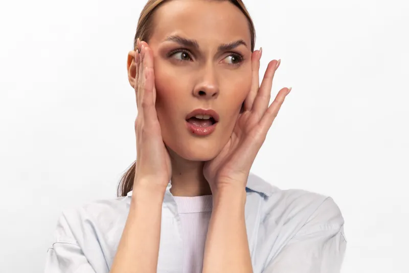 Surprised woman with light skin touches face with both hands, looking away on white background. Concept of shock, emotion, reaction, and beauty portrait with natural expression.