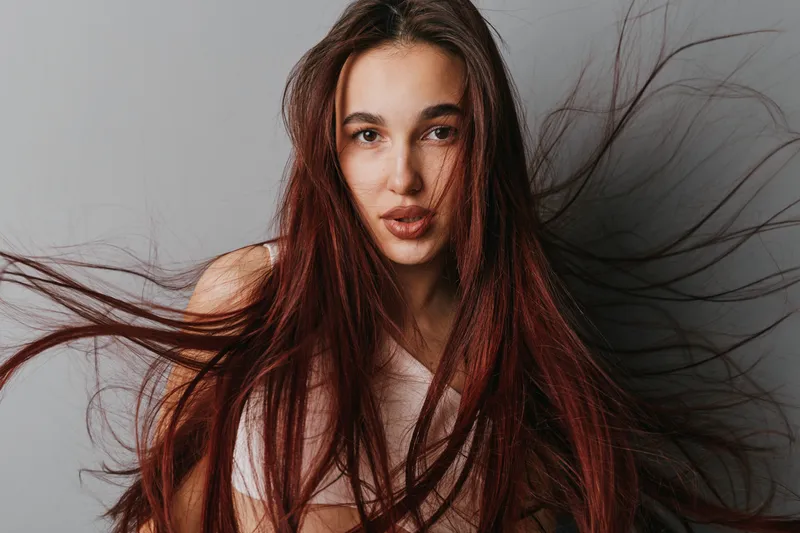 Woman with long brown hair looking at camera with parted lips and natural makeup on gray background. Portrait of young female with flowing hair and soft expression in studio light.