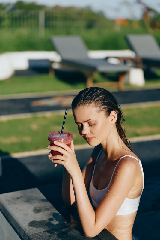 A young woman in a white bikini sits by a sunlit pool, sipping a pink smoothie with a striped straw, her expression calm and reflective in a carefree lifestyle moment.