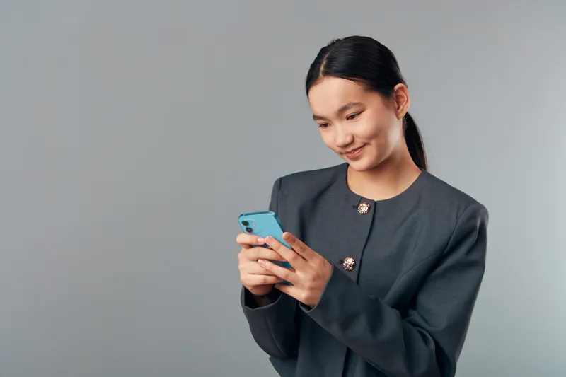 Young woman wearing dark blazer using blue smartphone with smile. Standing on gray background, texting and browsing with casual posture and focused expression in modern casual style.