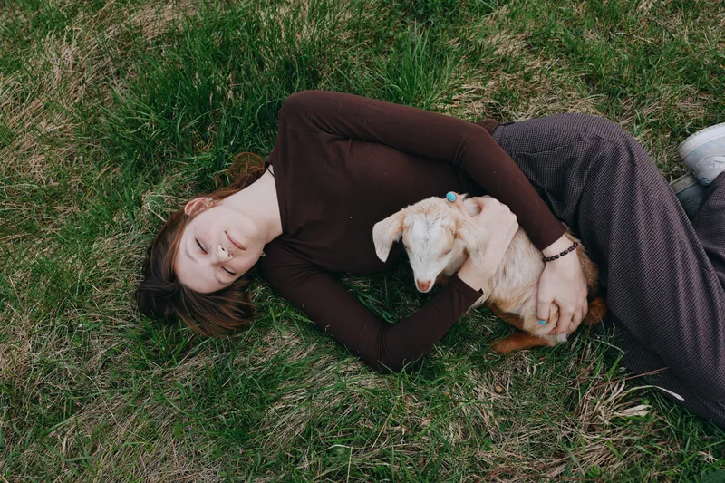 A young woman lies on grass holding a small baby goat close to her chest in a peaceful outdoor setting, capturing a tender moment of connection and calmness.