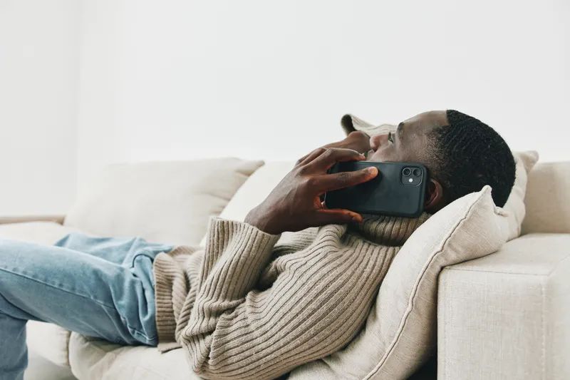 A young Black man in a cozy sweater reclining on a sofa while talking on a smartphone, creating a relaxed and modern atmosphere with soft neutral tones