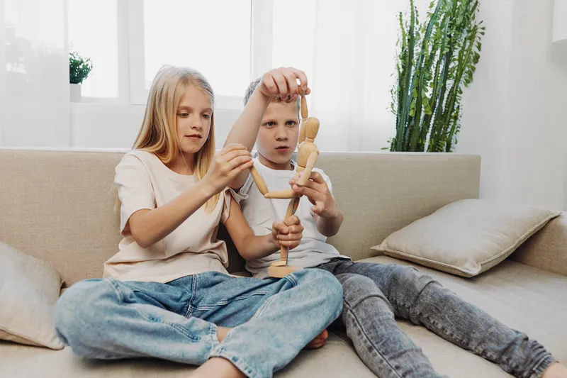 Two focused children sit on a sofa playing with a wooden artist mannequin in a bright living room with plants and cushions. Concept for creativity and childhood playtime.