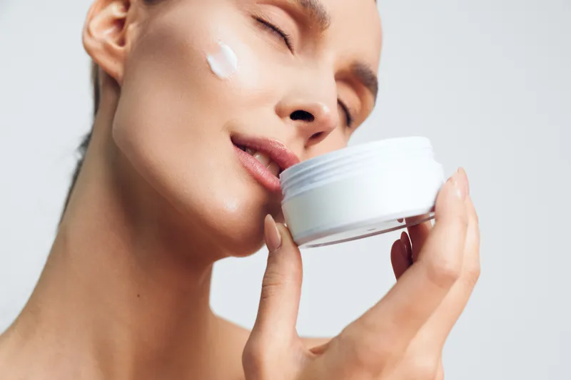 Young woman with clear skin applies moisturizing cream on face holds white jar close eyes enjoying skincare routine isolated on white background beauty concept.