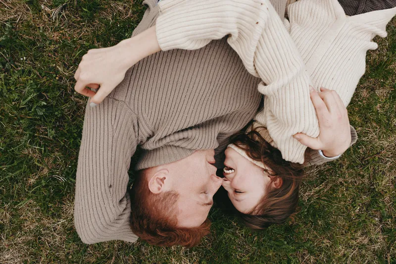 A couple lies on grass, smiling softly at each other in matching beige sweaters. A warm lifestyle moment outdoors, capturing affection, comfort, and relaxed togetherness in a candid, natural setting.