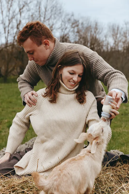 Young couple in cozy sweaters feed a baby goat with a bottle while sitting on hay in an outdoor natural setting during a calm autumn day.