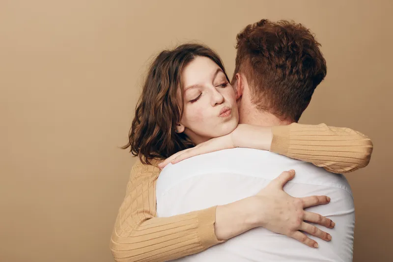 Young woman with curly hair hugs man wearing white shirt, eyes closed and peaceful expression. Intimate moment of affection and comfort on beige background. Concept of love and connection.