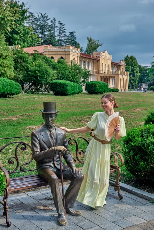 Woman in a pale yellow dress interacts with a bronze statue on a bench in a green park, with a historic building and trees in the background at dusk