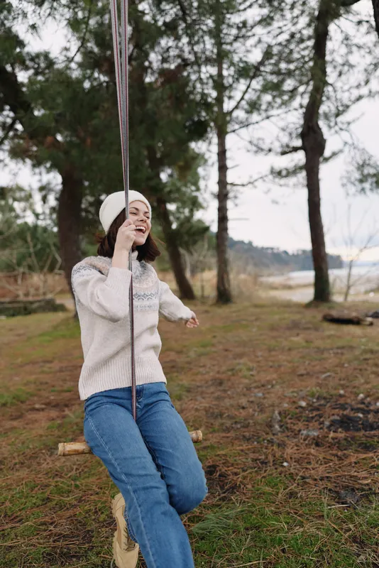woman on a wooden swing in outdoors nature, by a lake in a park, wearing sweater and beanie, enjoying a peaceful moment under trees