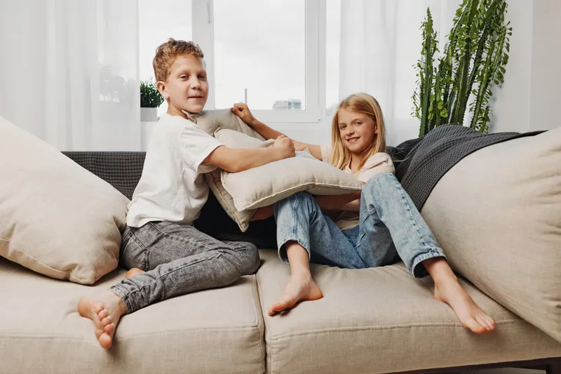 Two children playfully fight with pillows on a beige sofa in a bright living room with large windows and modern decor. The scene captures joyful family moments and childhood fun.