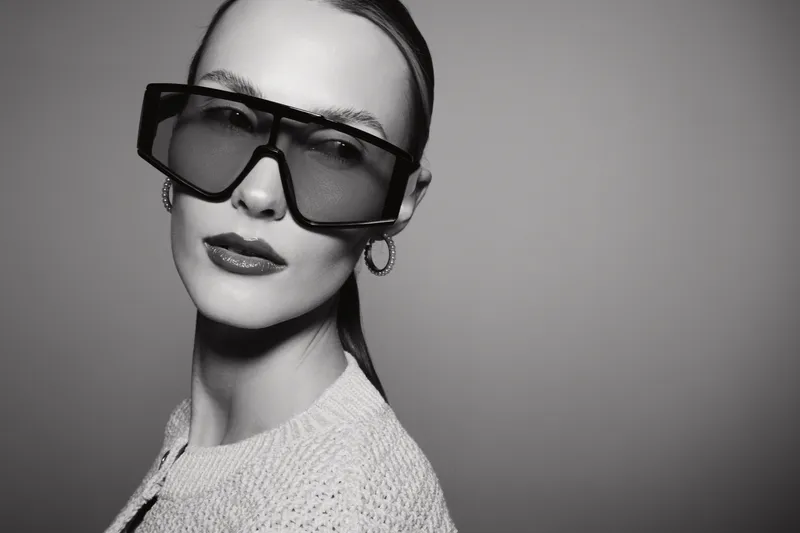 Stylish white woman with pale skin wearing large sunglasses and hoop earrings looks sideways. Black and white portrait in studio with soft lighting and minimalist background.