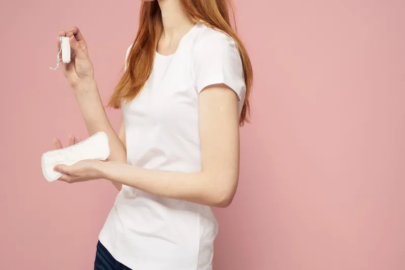 Женщина в джинсах и футболке держит в руке гигиеническую прокладку на розовом фоне