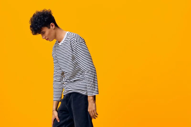 young man wearing striped shirt with curly hair posing thoughtfully on bright orange background, casual style and modern look, emotion of contemplation