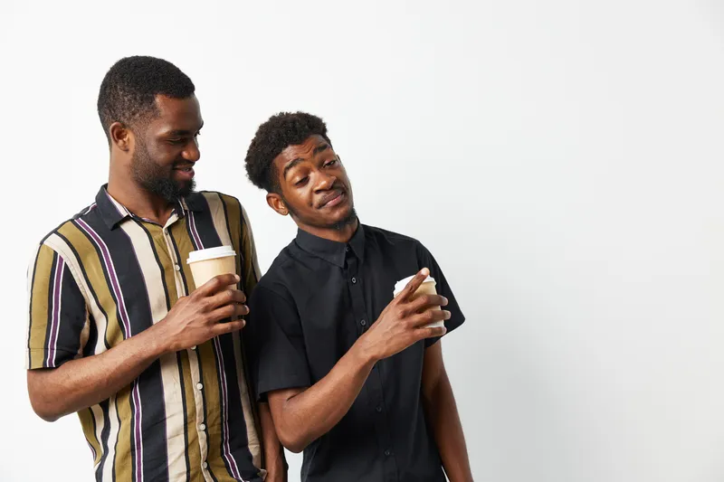 Two Black men sharing a light moment while holding coffee cups, showcasing friendship and laughter against a minimalistic white background.