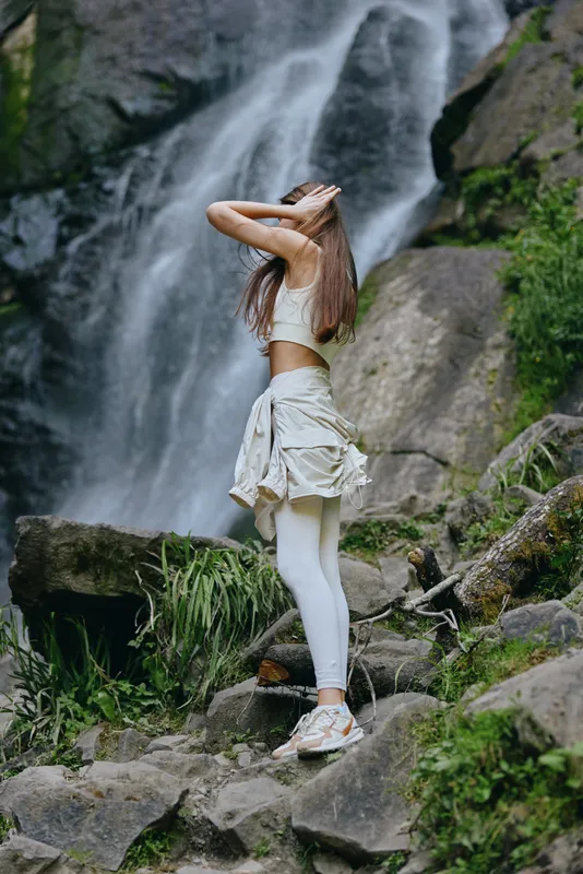 young woman standing near a rocky waterfall, wearing white activewear and sneakers, posing with hands on head in lush green natural environment, outdoor adventure scene