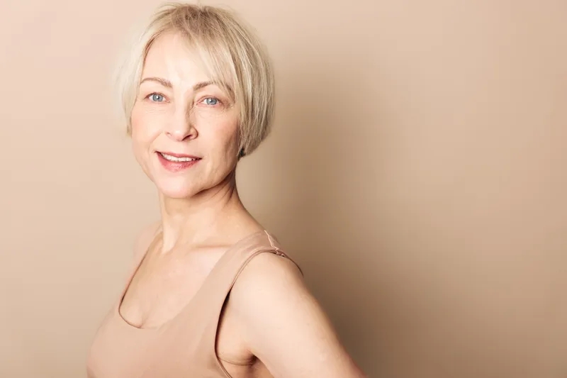 Smiling middle aged woman with short blonde hair wears beige sleeveless top, posing against soft beige background. Concept of confidence, natural beauty, and positive aging.