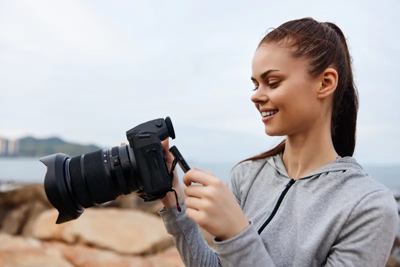 Young smiling photographer adjusting camera on rocky beach, casual sportswear, capturing creativity by the sea, vibrant emotions