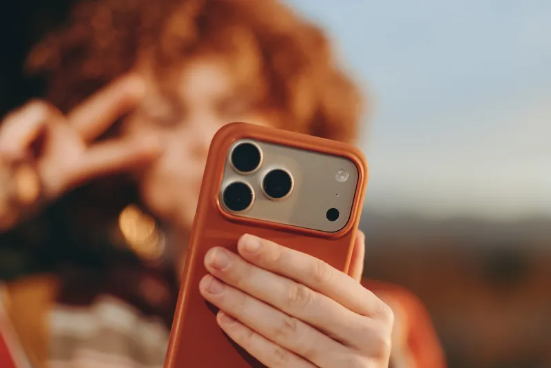 Woman holds a smartphone for a selfie outdoors in autumn, closeup of her hand with an orange phone case, blurred portrait background, warm light, lifestyle moment showing technology and communication.