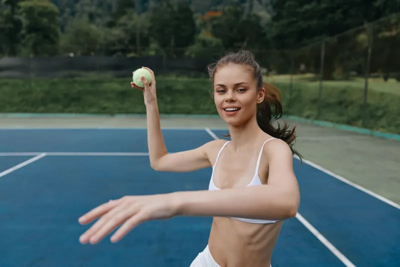 Young woman in sportswear throwing a tennis ball on a blue court with a joyful expression, highlighting an active lifestyle and the essence of sports