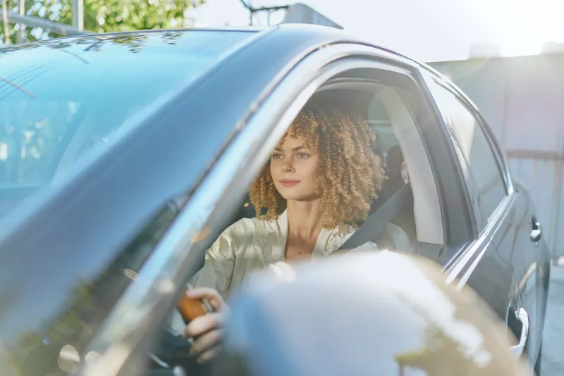 young woman driving car outdoors on sunny day with casual clothes and curly hair, focused and calm, bright natural light in urban setting