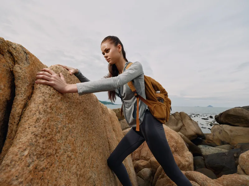 Young woman climbing rocky terrain with backpack, wearing grey sportswear, showcasing determination, with seaside background and muted colors, perfect for adventure themes