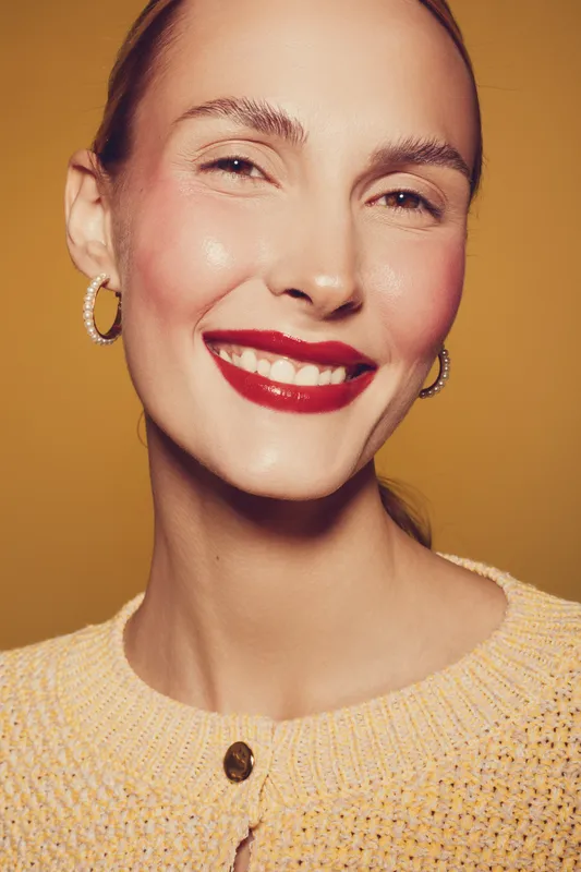 Smiling woman with red lipstick and hoop earrings wears yellow knitted sweater. Close-up portrait on warm orange background. Concept of beauty, fashion, happiness, and confident female model.
