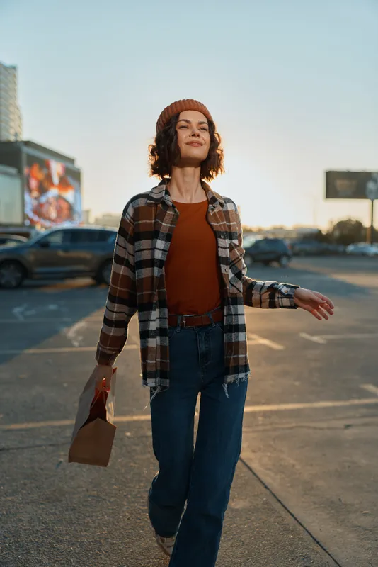 Woman walking in urban parking lot wearing beanie and plaid shirt, holding takeout bag, joyful expression at sunset with candid lifestyle, authenticity, golden hour glow and mindful living.