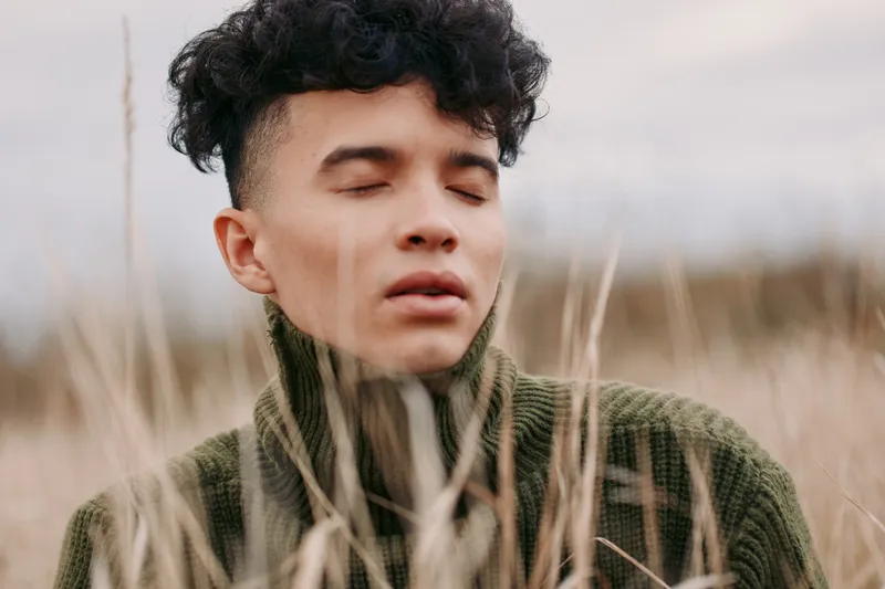 A young man in a heavy knitted sweater closes his eyes amid tall dry grasses, conveying an authentic and truthful moment of quiet introspection and a natural, unposed outdoor portrait mood.