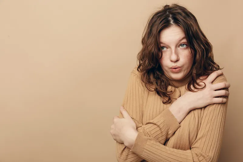 Young woman in beige sweater hugs herself with crossed arms, expressing cold or discomfort. Neutral background and casual style create a simple, relatable concept of feeling chilly or vulnerable.
