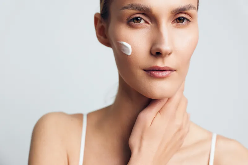 Young woman with clear skin applying moisturizing cream on cheek, natural look, soft lighting, neutral background, gentle skincare routine, calm expression
